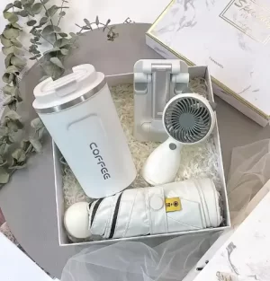 white-everyday-comfort-gift-set.jpg Minimalist white gift box containing a travel coffee mug, compact umbrella, handheld cat-ear mini fan, and a phone stand set against marble packaging.