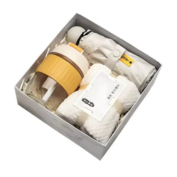 Cozy lifestyle gift set containing a yellow glass tumbler with straw, a folded white UPF 50+ umbrella, and a rolled plush towel arranged in a gift box.