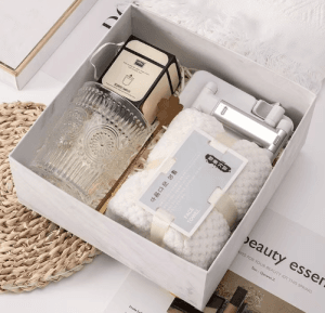 Cozy self-care luxury gift box containing an embossed glass tumbler, boxed scented candle, white waffle face towel, and silver phone stand in a marble presentation box.