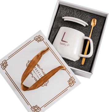 Personalised white ceramic mug with custom letter monogram, ceramic lid, and gold spoon inside a luxury presentation gift box with ribbon handles.