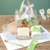 Daisy Clear Tote Spa & Self-Care Gift Bag A transparent tote bag decorated with white daisy prints and a ribbon tied with "JUST FOR YOU" text on beige handles. The tote contains self-care items alongside a rolled white plush towel, a mini perfume bottle with a red bow, a ribbed glass tumbler, a gold flower spoon, and white box packaging set on top of printed paper.