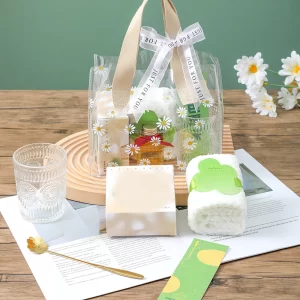 A transparent tote bag decorated with white daisy prints and a ribbon tied with "JUST FOR YOU" text on beige handles. The tote contains self-care items alongside a rolled white plush towel, a mini perfume bottle with a red bow, a ribbed glass tumbler, a gold flower spoon, and white box packaging set on top of printed paper.