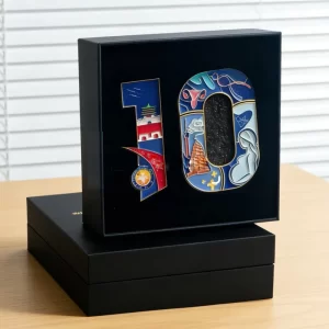A luxury black velvet-lined presentation box holding a custom multi-panel crest enamel pin featuring detailed architectural and health illustrations for corporate milestone celebrations.