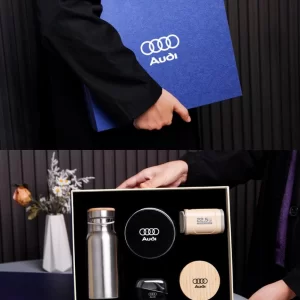 Two-part composite image showing a person holding a textured blue Audi branded gift box on top, and opening the box below to reveal a stainless steel water bottle, a wireless charging pad, a 22.5W power bank, wireless earbuds in a charging case, and a wooden coaster nestled in custom black foam.